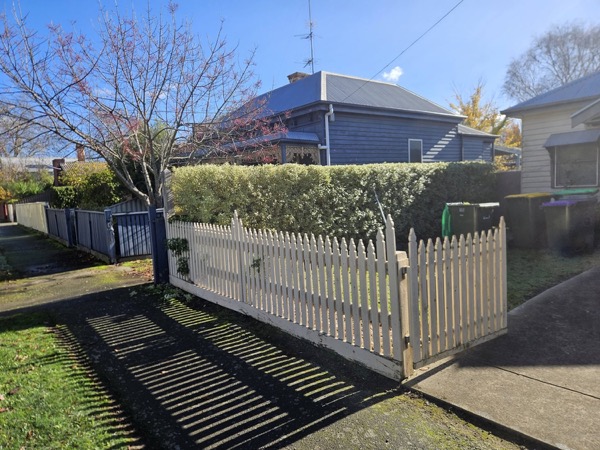 A clipped hedge and picket gate in front of a weatherboard cottage