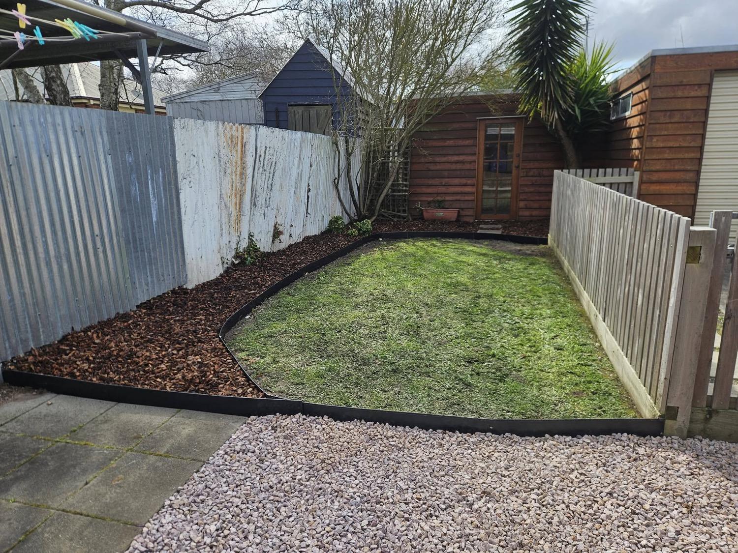 A back lawn with a curved mulched garden bed and a pebbled strip along the fence